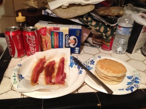 Photo of a paper plate with 3 bacon strips next to a paper plate with a stack of pancakes with a dinner knife connecting the two. There are assorted food items in the background.
